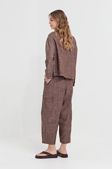 A woman stands in profile wearing a brown and taupe plaid two-piece linen set consisting of a cropped long-sleeve top and wide-leg cropped pants by Verge, paired with black sandals, against a plain white studio background.