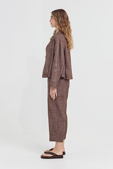 A woman stands in profile wearing a matching brown plaid two-piece outfit consisting of a cropped long-sleeve blazer and cropped trousers by Verge, paired with dark brown flat sandals, against a plain white studio background.