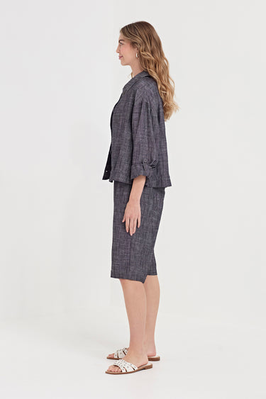 A woman stands in profile wearing a matching gray plaid two-piece set consisting of a cropped jacket with three-quarter sleeves and a knee-length skirt by Verge, paired with white slide sandals, against a plain white studio background.