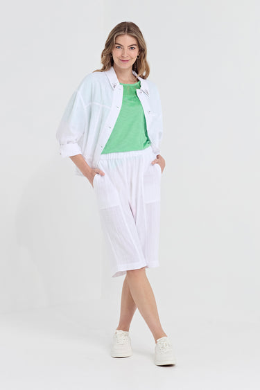 A woman modeling a white lightweight button-up shirt over a bright green crewneck top, paired with white shorts by Verge and white sneakers, against a plain white studio background.