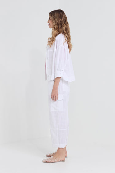A woman stands in profile wearing a white two-piece lounge set consisting of a cropped button-front shirt and matching wide-leg pants with patch pockets by Verge, paired with clear slip-on mules, against a plain white studio background.