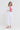 A woman modeling a white linen-blend two-piece set consisting of a cropped button-up shirt with a tie waist and matching relaxed-fit pants by Verge, worn over a pink top.