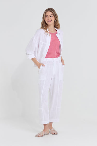 A woman modeling a white linen two-piece set consisting of a button-up shirt and matching wide-leg cropped pants, worn over a pink t-shirt by Verge and paired with pink patterned flat sandals, on a light gray studio background.