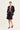 A model stands against a plain white studio background wearing a black tailored blazer and matching black shorts over a colorful paisley-patterned top by Verge, with black lace-up flat shoes.