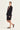 A woman models a black tailored set consisting of a blazer with decorative silver studs on the shoulders and matching shorts by Verge, paired with a patterned top and black strappy mule flats, against a plain off-white studio background.