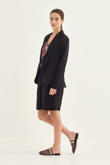 A woman models a black tailored set consisting of a blazer with decorative silver studs on the shoulders and matching shorts by Verge, paired with a patterned top and black strappy mule flats, against a plain off-white studio background.