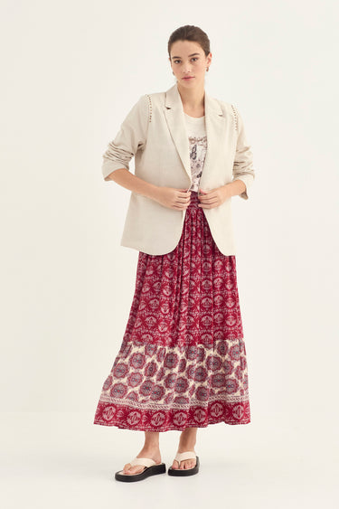 A woman modeling a long-sleeve cream blazer by Verge over a white top and a long burgundy maxi skirt with a cream bohemian pattern and border, paired with black slide sandals, against a plain off-white studio background.