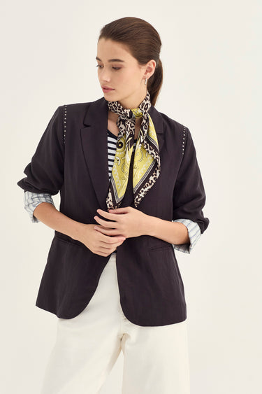 A woman models a dark navy blazer with decorative silver studs along the shoulders, styled over a striped shirt and a yellow-and-black patterned silk scarf, paired with white trousers by Verge, against a plain off-white background.