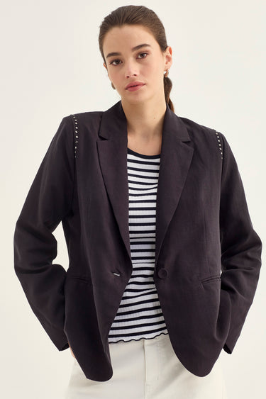 A woman models a dark navy blazer with decorative silver studs along the shoulders, styled over a striped shirt paired with white trousers by Verge, against a plain off-white background.