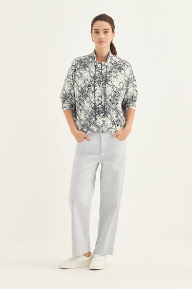 A woman stands against a plain white studio background wearing a black and white floral print button-up shirt with rolled sleeves, light gray wide-leg trousers by Verge, and white sneakers.