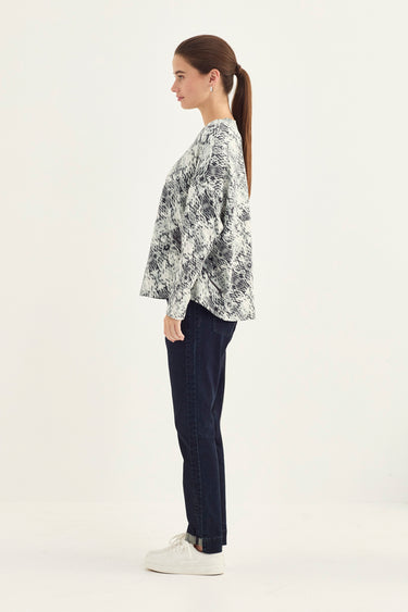 A woman in profile wearing a snake print jacket pattern long-sleeve top, dark navy jeans by Verge, and white sneakers, standing against a plain off-white studio background.