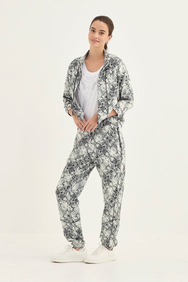 A woman modeling a matching two-piece lounge set featuring a snake print jacket pattern, consisting of a lightweight zip-up jacket and relaxed pants, styled with a white t-shirt by Verge and white sneakers against a plain white studio background.