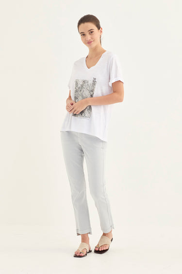 A woman modeling a white V-neck short-sleeve t-shirt with a black and gray graphic print on the front, paired with light gray cropped pants by Verge and flat sandals, against a plain white studio background.