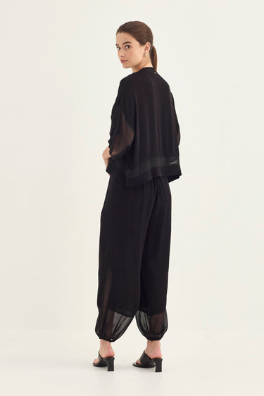 A model wearing a black sheer two-piece outfit consisting of a short-sleeve top and matching wide-leg pants with gathered sheer cuffs by Verge, shown from a rear three-quarter angle against a plain white studio background.