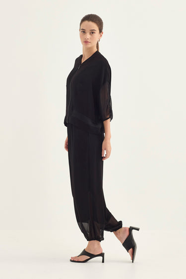 A woman modeling a black sheer long-sleeve top and matching black wide-leg trousers, paired with black heeled sandals by Verge, against a plain white studio background.