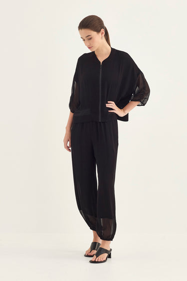 A woman modeling a black two-piece outfit consisting of a short-sleeve cropped jacket with sheer three-quarter sleeves and matching loose wide-leg pants, paired with black sandals by Verge, against a plain white studio background.