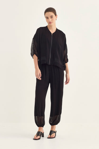 A model wearing a black two-piece outfit consisting of a cropped zip-up jacket with sheer sleeves and matching loose trousers with sheer ankle cuffs, paired with black heeled sandals by Verge, against a plain off-white studio background.