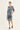 A woman modeling a blue and white patterned patchwork shirt dress by Verge with three-quarter sleeves and front pockets, paired with black sandals, against a plain light background.
