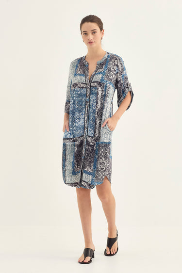 A woman modeling a blue and white patterned patchwork shirt dress by Verge with three-quarter sleeves and front pockets, paired with black sandals, against a plain light background.