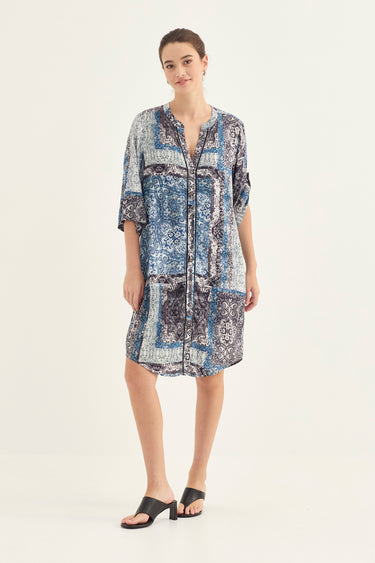 A woman modeling a short-sleeve button-front tunic dress by Verge with an abstract blue, white, and gray pattern, paired with black slide sandals, against a plain light background.