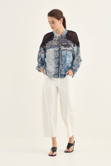 A woman modeling a long-sleeve button-up blouse with a blue and white paisley pattern, black mesh shoulder panels, and white cropped pants by Verge , standing against a plain light background.