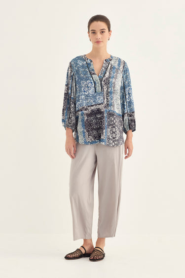 A woman modeling a blue and gray patterned blouse with three-quarter sleeves, paired with light beige wide-leg cropped pants by Verge and black woven sandals, against a plain off-white studio background.