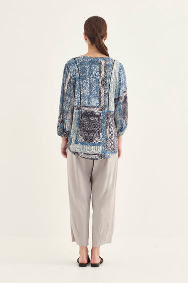 A woman viewed from the back wearing a loose long-sleeve patchwork blouse in shades of blue, gray, and white, paired with light beige cropped pants by Verge and black sandals, against a plain off-white studio background.