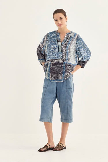 A woman modeling a blue and white patterned blouse with voluminous sleeves, paired with light blue cropped denim shorts by Verge and dark flat sandals, against a plain off-white studio background.