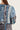 Close-up back view of a woman wearing a blue, white, and black paisley patchwork blouse by Verge against a light beige background.