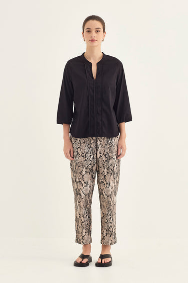 A woman stands against a plain light background wearing a black three-quarter sleeve top with a split neckline and beige-and-brown snakeskin-print cropped pants by Verge, paired with black sandals.