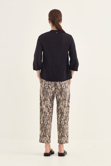 A woman modeling viewed from the back wearing a black long-sleeve top and beige-and-brown snakeskin print pants by Verge, paired with black sandals, against a plain off-white studio background.