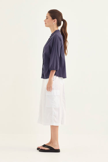 A woman modeling a denim three-quarter sleeve top and white shirt by Verge, paired with black flip-flops, against a plain white studio background.