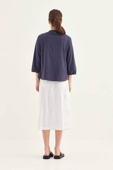 A woman viewed from the back wearing a navy blue cropped three-quarter sleeve top and a white midi skirt by Verge, standing against a plain off-white studio background.