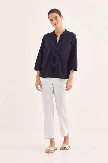A woman modeling a dark navy blue linen shirt with three-quarter sleeves and a split neckline, paired with white cropped trousers by Verge and flat sandals, against a plain off-white studio background.