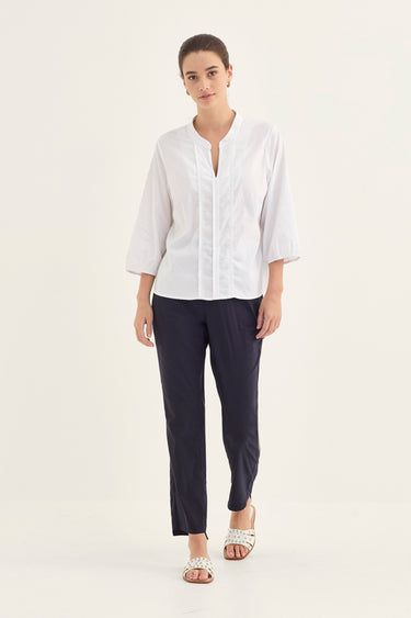 A woman modeling a white three-quarter sleeve button-up blouse with a split neckline, paired with dark navy trousers by Verge and white heeled sandals, against a plain off-white studio background.