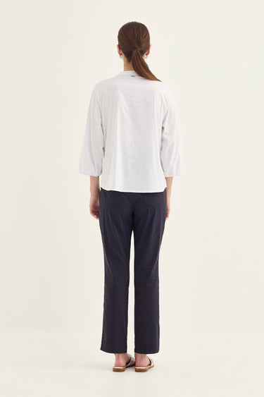 A woman shown from the back wearing a white three-quarter sleeve top and dark navy pants by Verge, standing against a plain off-white studio background.