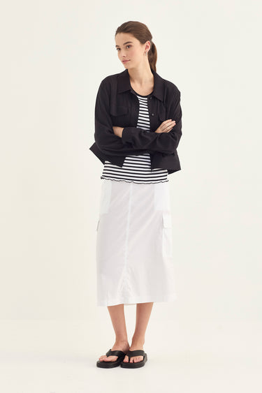 A woman modeling a black cropped button-up shirt over a black and white striped top, paired with a white midi skirt by Verge and black sandals, against a plain off-white studio background.