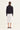 A woman viewed from behind wearing a black, cropped, long-sleeve jacket over a white midi skirt by Verge, paired with black flat shoes, against a plain off-white studio background.