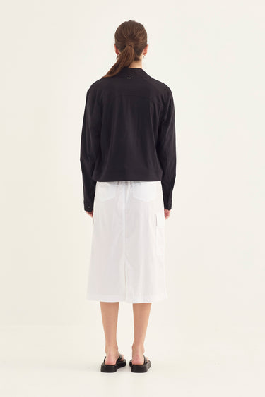 A woman viewed from behind wearing a black, cropped, long-sleeve jacket over a white midi skirt by Verge, paired with black flat shoes, against a plain off-white studio background.