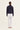 A woman viewed from the back wearing a dark navy long-sleeve cropped jacket, white trousers by Verge, and flat sandals, standing against a plain off-white studio background.