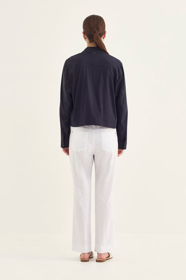 A woman viewed from the back wearing a dark navy long-sleeve cropped jacket, white trousers by Verge, and flat sandals, standing against a plain off-white studio background.