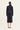 A model viewed from the back wearing a dark navy blue two-piece outfit consisting of a cropped long-sleeve jacket and a matching midi skirt by Verge, paired with white heeled sandals, against a plain off-white studio background.