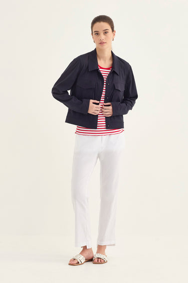 A woman modeling a dark navy utility jacket over a red and white striped t-shirt, paired with white cropped pants by Verge and white slide sandals, against a plain light background.