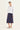 A woman modeling a white long-sleeve button-up jacket and a dark navy midi skirt by Verge, paired with white slide sandals, against a plain off-white studio background.