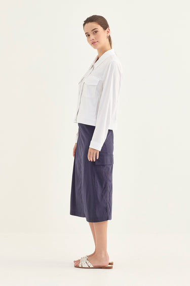 A woman stands in profile wearing a white long-sleeve button-up shirt and a dark navy midi skirt by Verge with large patch pockets, paired with white slide sandals, against a plain off-white studio background.