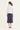 A woman is shown from behind wearing a white cropped long-sleeve shirt and a dark slate blue midi skirt by Verge, standing against a plain off-white studio background.