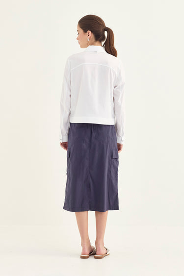 A woman is shown from behind wearing a white cropped long-sleeve shirt and a dark slate blue midi skirt by Verge, standing against a plain off-white studio background.