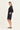 A woman modeling a loose, short black dress by Verge with three-quarter sleeves and side pockets, standing in profile against a plain off-white studio background.