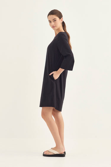 A woman modeling a loose, short black dress by Verge with three-quarter sleeves and side pockets, standing in profile against a plain off-white studio background.