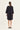 A woman viewed from the back wearing a loose black dress by Verge with three-quarter sleeves and black flat shoes, standing against a plain light background.